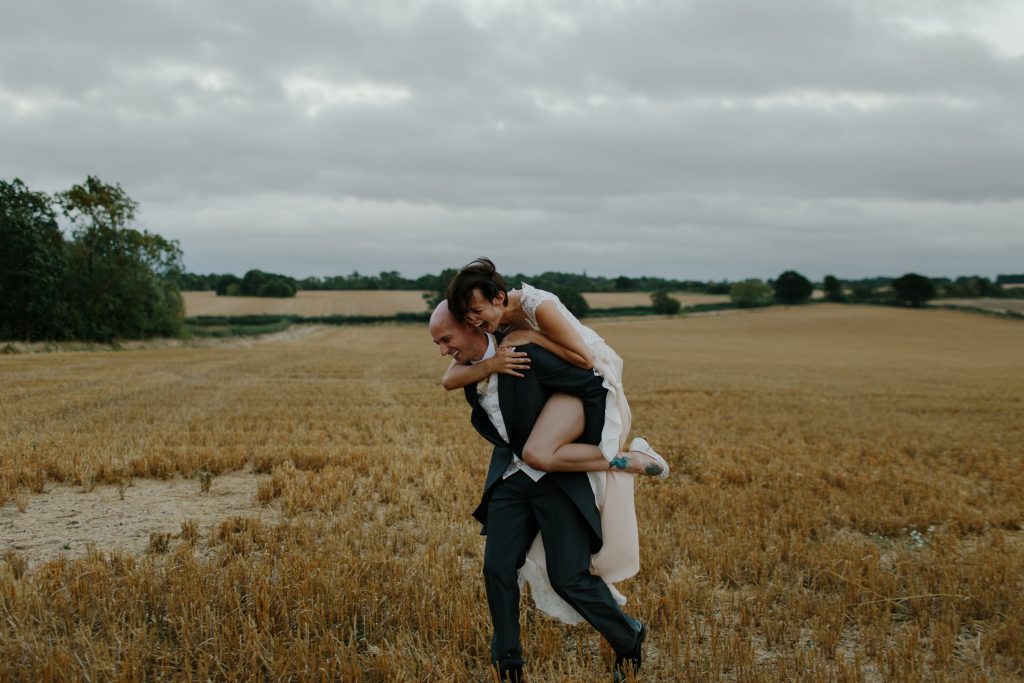 bride rides groom piggy back through farm field