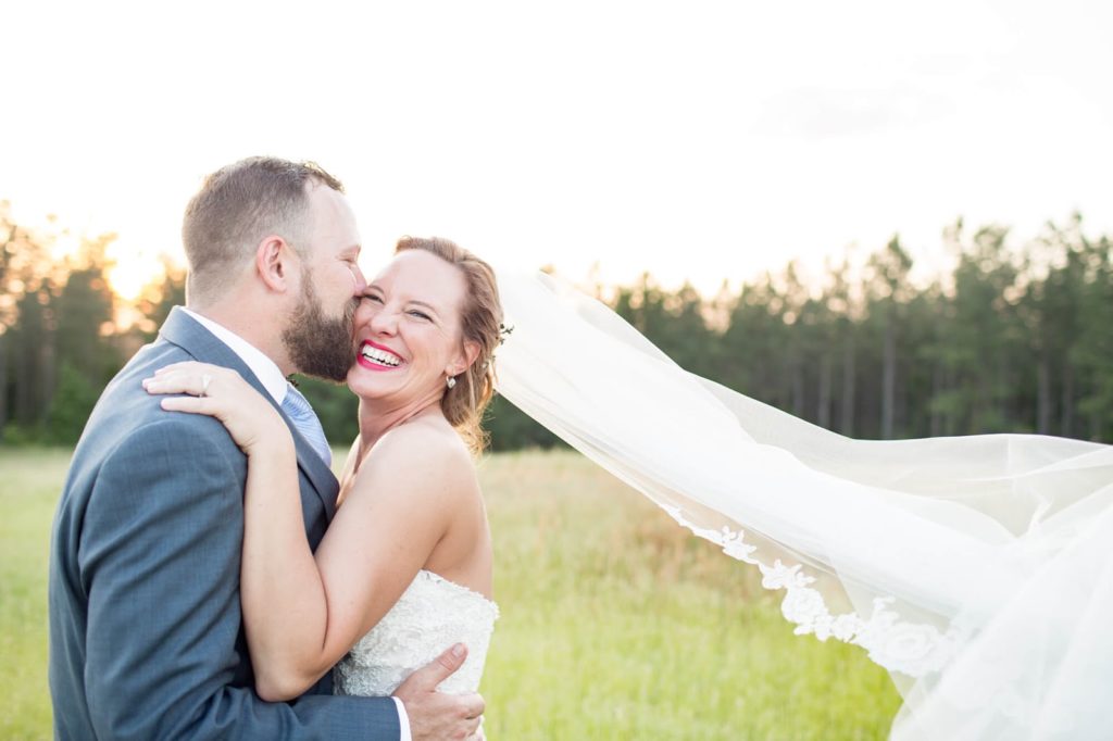 groom kisses his bride in a sunny meadow as veil gently flutters in the breeze