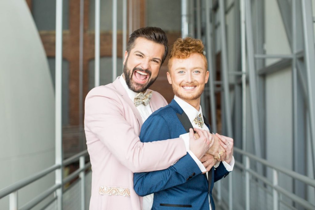 two grooms embrace smiling on their wedding day, pink suit and blue suit