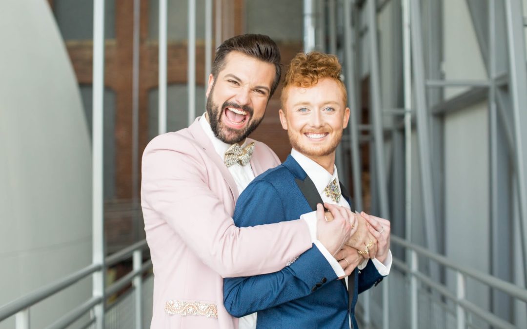 two grooms embrace smiling on their wedding day, pink suit and blue suit