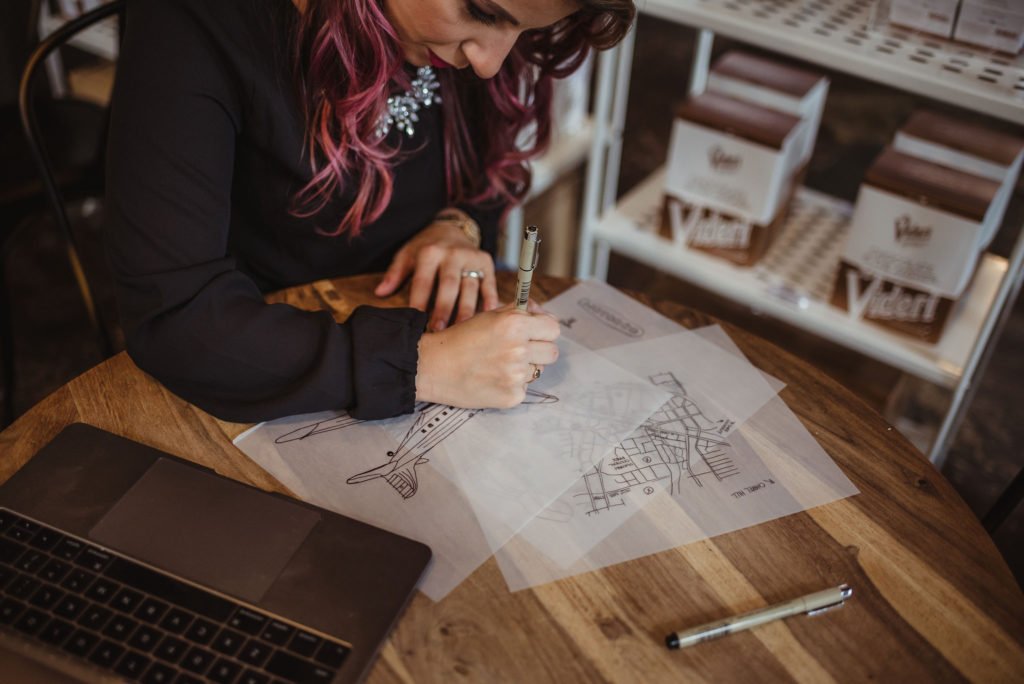 Kasey sits inside a coffee shop sketching images of planes, and maps for a custom commissioned piece
